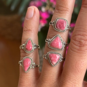 May include: A collection of silver rings with pink gemstones in various shapes: teardrop, triangle, and square. Each ring features a detailed silver band with a twisted rope design around the gemstone. The rings are displayed on a hand.