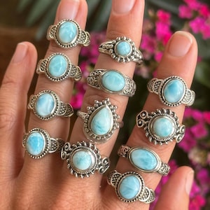 Könnte beinhalten: Eine Sammlung von Silberringen, jeder mit einem hellblauen, ovalen oder tropfenförmigen Edelstein. Die Ringe werden auf einer Hand präsentiert, die verschiedene filigrane Silberbanddesigns zeigt. Der Hintergrund umfasst grüne Blätter und rosa Blüten.