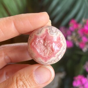 May include: A spherical gemstone, displaying a pink and white banded design. The stone is held in a hand, with a blurred backdrop of green foliage and vibrant pink flowers.