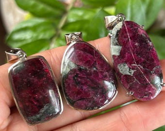 Puede incluir: Tres colgantes con rodajas de piedras preciosas de color magenta intenso y verde oscuro pulidas, engastadas en marcos de metal plateado. Los colgantes tienen formas rectangular, de gota y ovalada, cada uno con una anilla metálica en la parte superior.
