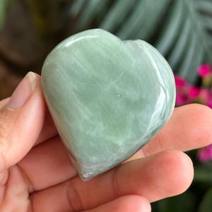 May include: A polished, heart-shaped stone in a pale, translucent sage green color with subtle white veining throughout the surface. The stone has a smooth, glossy finish and a rounded, symmetrical shape that tapers to a point at the bottom.