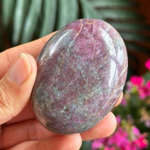 May include: A smooth, polished oval-shaped stone featuring a mottled blend of deep magenta, dark purple, and muted teal-green tones. The surface displays natural crystalline inclusions and fine white veining throughout the rock's dense texture.