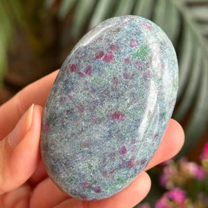 Pode incluir: Uma pedra preciosa polida de forma oval e lisa, com uma textura manchada sobre uma base de tons azul-acinzentados suaves, intercalada com inclusões vibrantes de rosa magenta e pequenas manchas verde-escuras na superfície.