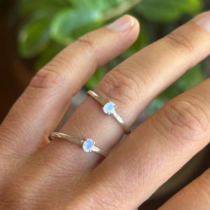 May include: Two thin silver rings, each featuring a small, oval-shaped, iridescent moonstone set in a simple metal band. The stones display a soft, pale blue glow, and the rings are worn on two fingers, showcasing the polished silver finish.