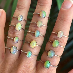 May include: A collection of twelve thin silver rings, each featuring a single small oval-shaped opal gemstone. The opals display iridescent flashes of green, blue, and yellow light. Each ring has a simple, slender metal band and a prong setting.