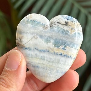 以下が含まれることがあります： 白、水色、濃い青の水平な帯模様が入った、磨き上げられたハート型の石。石の表面全体には自然な鉱物のインクルージョンや筋が見られ、滑らかで丸みを帯びた形状の中に多様な色彩が広がっています。