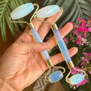 Puede incluir: Dos rodillos faciales de jade con mangos dorados y piedras ovaladas de color azul claro. Los rodillos se sostienen en una mano, con un fondo de hojas verdes y flores rosas.