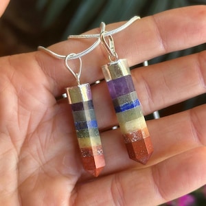 Könnte beinhalten: Zwei Chakra-Anhänger-Halsketten. Jeder Anhänger ist ein vertikales, rechteckiges Prisma mit Schichten aus roten, orangen, gelben, grünen, blauen, indigo- und violetten Steinen. Die Anhänger sind an einer silbernen Kette aufgehängt.
