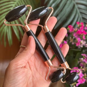 Puede incluir: Dos rodillos faciales negros con marcos de metal color oro rosa. Los rodillos se sostienen en una mano. El fondo incluye plantas verdes y flores rosas. Aspecto liso y pulido.