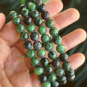 Pode incluir: Close-up de várias fileiras de pulseiras de contas verdes e vermelho escuro. As contas redondas têm uma aparência salpicada. As pulseiras são seguradas em uma mão, com um fundo verde desfocado.