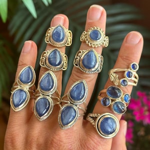 Pode incluir: Uma coleção de anéis de prata com pedras preciosas de cianite azul em várias formas, incluindo cortes em gota, ovais e redondos. Os anéis exibem um trabalho de metal intrincado, com alguns designs apresentando várias pedras numa única banda.