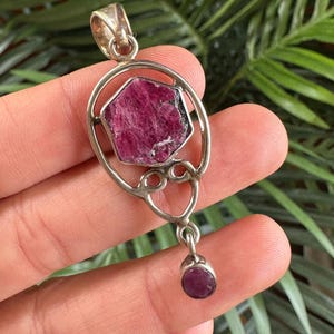 Puede incluir: Un colgante de plata con una gran piedra preciosa en bruto pentagonal de color rosa rojizo intenso engastada en un marco circular con volutas decorativas. Una piedra más pequeña, redonda y de color rosa rojizo intenso cuelga de la base.