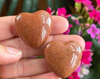 May include: Two polished heart-shaped goldstone pieces with a smooth, reflective surface. The stones feature a deep copper-brown color with embedded metallic glitter particles that catch the light, creating a sparkling, starry appearance.