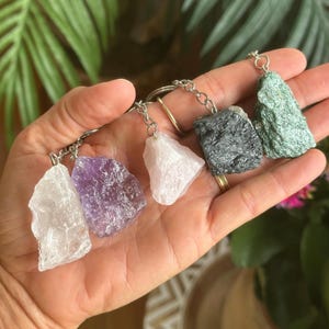 May include: A hand displays six raw crystal keychains in various colors: clear, purple, white, black, and green. Each crystal is attached to a silver chain and keyring. The background features green foliage and a hint of pink flowers.