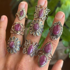 May include: A collection of seven silver rings, each featuring a polished, oval-shaped magenta gemstone. The designs include butterfly motifs, floral-inspired settings with scalloped edges, and geometric bands, all showcasing intricate metalwork details.