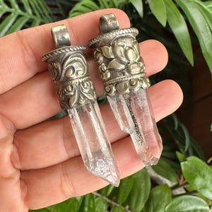 Pode incluir: Dois pingentes de cristal de quartzo transparente e pontiagudo com tampas de metal prateado adornadas com gravuras florais e volutas. Cada pingente inclui uma argola de metal resistente no topo e exibe inclusões naturais na pedra.