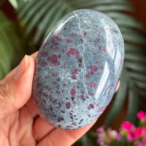 May include: A polished, oval-shaped stone featuring a smooth, glossy surface with a mottled blue-grey base color and numerous embedded deep magenta and dark red crystalline inclusions scattered throughout the entire face of the mineral specimen.