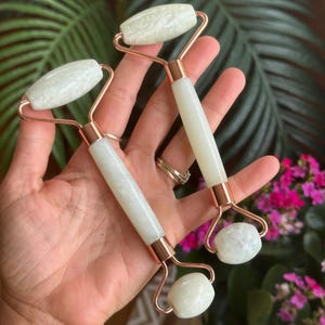 Puede incluir: Dos rodillos faciales de jade con cabezales y mangos de piedra blanca, engarzados en un marco de metal color cobre. Los rodillos se sostienen en una mano, con un fondo de hojas verdes y flores rosas.