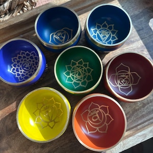 Może przedstawiać: Zestaw siedmiu kolorowych misek śpiewających. Każda miska jest pomalowana na inny kolor, w tym niebieski, zielony, żółty i czerwony. Każda miska ma złoty symbol czakry na środku i złoty pasek na obrzeżu. Miski są ułożone na drewnianej tacy.