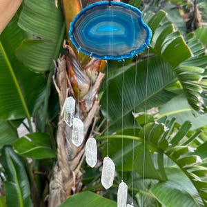 May include: A blue and white agate slice wind chime with clear quartz crystal pendants. The agate slice is oval-shaped with a polished surface. The crystals are suspended by thin, clear strings, creating a decorative and natural home decor item.
