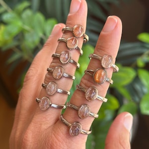 May include: A collection of ten silver rings, each featuring a single oval-shaped sunstone cabochon. The stones exhibit varying shades of orange, peach, and translucent white with internal inclusions, set in simple metal bands worn on fingers.