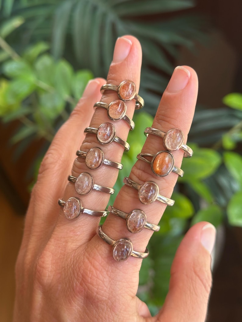 May include: A collection of ten silver rings, each featuring a single oval-shaped sunstone cabochon. The stones exhibit varying shades of orange, peach, and translucent white with internal inclusions, set in simple metal bands worn on fingers.