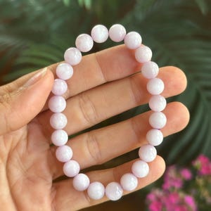 K&ouml;nnte beinhalten: Ein helllila Perlenarmband. Das Armband besteht aus runden, polierten Perlen. Die Perlen haben eine weiche, pastellviolette Farbe. Das Armband wird vor einem verschwommenen gr&uuml;nen und rosa Hintergrund gezeigt.