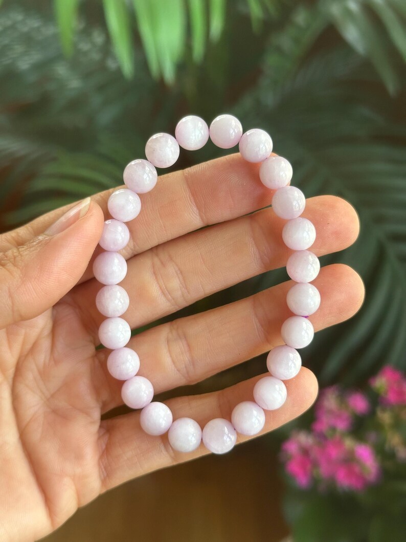 K&ouml;nnte beinhalten: Ein helllila Perlenarmband. Das Armband besteht aus runden, polierten Perlen. Die Perlen haben eine weiche, pastellviolette Farbe. Das Armband wird vor einem verschwommenen gr&uuml;nen und rosa Hintergrund gezeigt.
