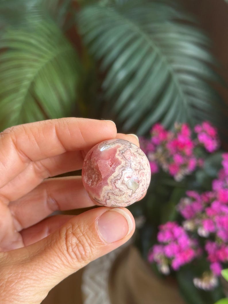 May include: A spherical gemstone, polished to a high sheen, displaying a swirling pattern of pink and cream hues. The stone is held in a hand, with a backdrop of green foliage and bright pink flowers.