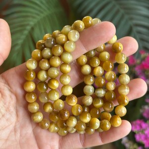 May include: Several strands of golden tiger eye gemstone bracelets. The beads are round and have a polished, reflective surface. The bracelets are held in a hand, with a blurred background of green foliage and pink flowers.