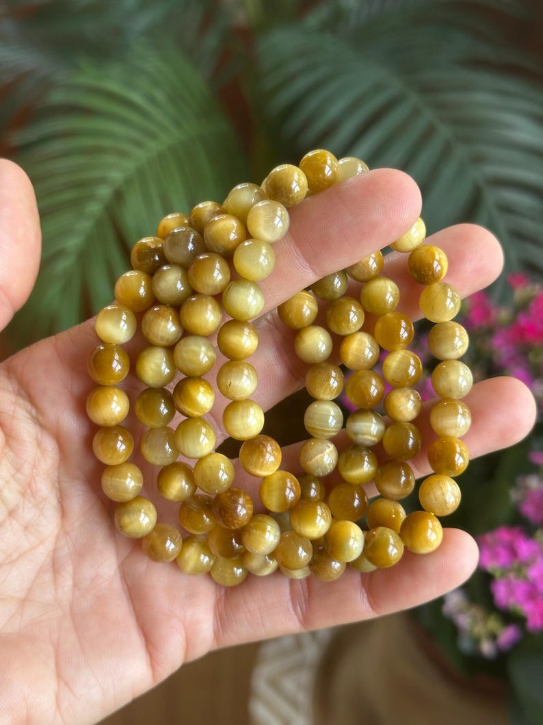 May include: Several strands of golden tiger eye gemstone bracelets. The beads are round and have a polished, reflective surface. The bracelets are held in a hand, with a blurred background of green foliage and pink flowers.