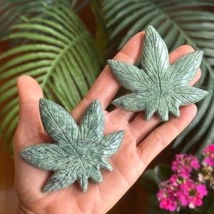Peut inclure: Deux sculptures en forme de feuilles de cannabis en pierre verte sculptée. Les feuilles présentent des veines détaillées et une surface texturée. Les sculptures sont tenues dans une main, avec un fond de feuillage vert et de fleurs roses.