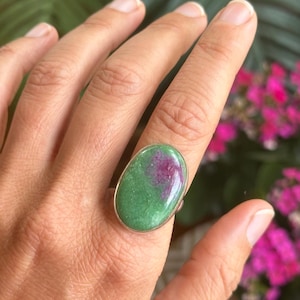 May include: A silver ring featuring a large, oval-shaped gemstone. The stone is a mix of green and magenta hues, set in a polished silver band. The ring is displayed on a hand, with a blurred background of green foliage and pink flowers.