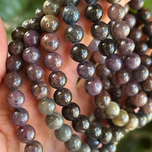 May include: A multi-strand beaded bracelet featuring polished, spherical stones in shades of deep purple, charcoal gray, and muted olive green. The beads are strung together in a circular arrangement, showcasing natural variations in color and texture.