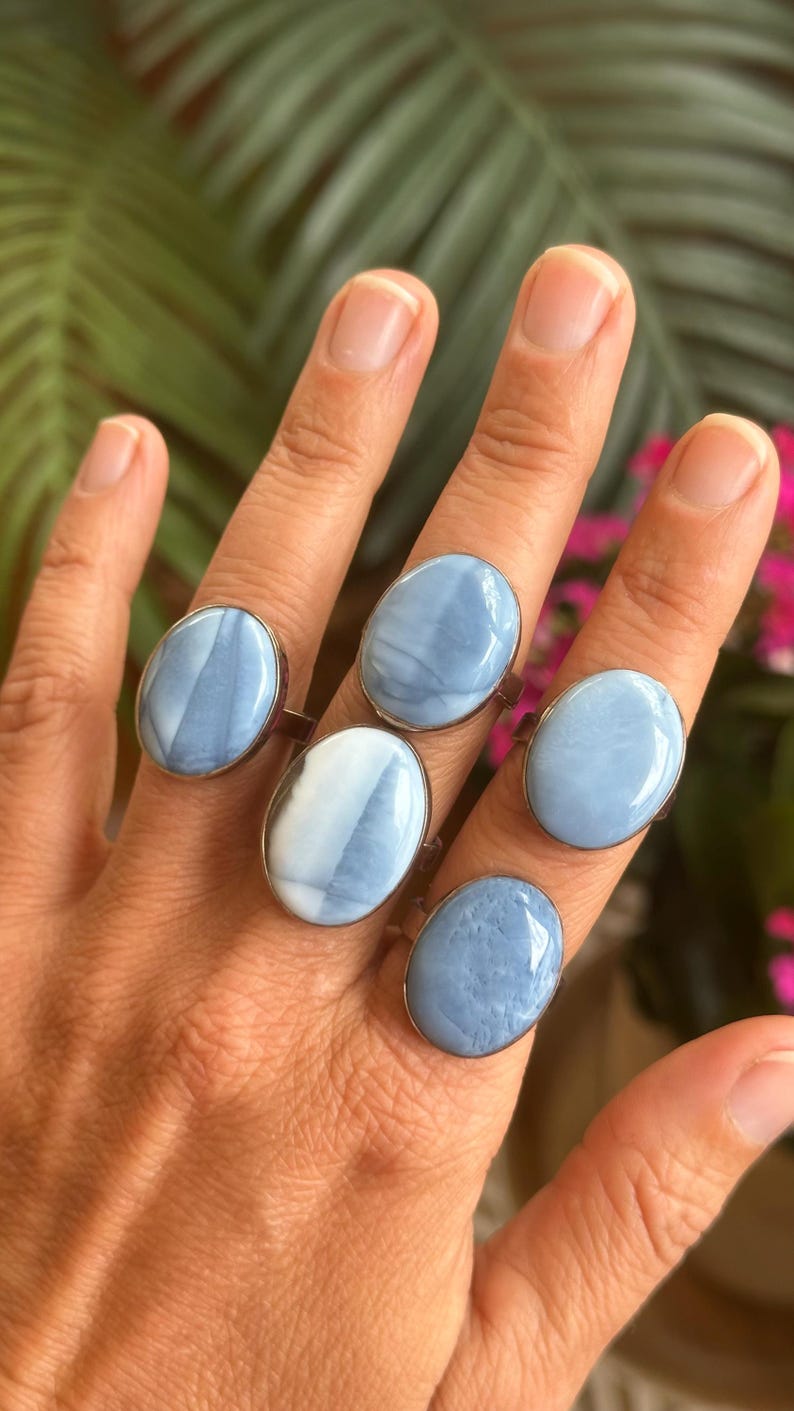 May include: Five oval-shaped rings with light blue and white stone settings are displayed on a hand. The rings have a silver-colored band. The background features green foliage and pink flowers.