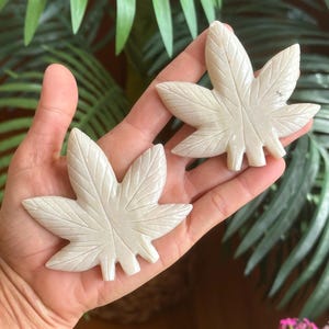 May include: Two carved, off-white stone cannabis leaf sculptures. The leaves are held in a hand, with detailed veining and a naturalistic design. The background features green foliage and pink flowers.