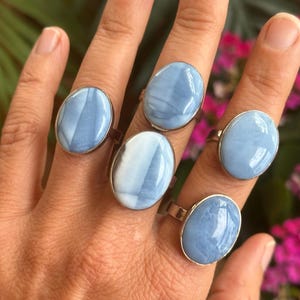 May include: Five silver rings with oval, light blue gemstones are displayed on a hand. The gemstones have a marbled appearance with white and blue swirls. The rings are set on a silver band.