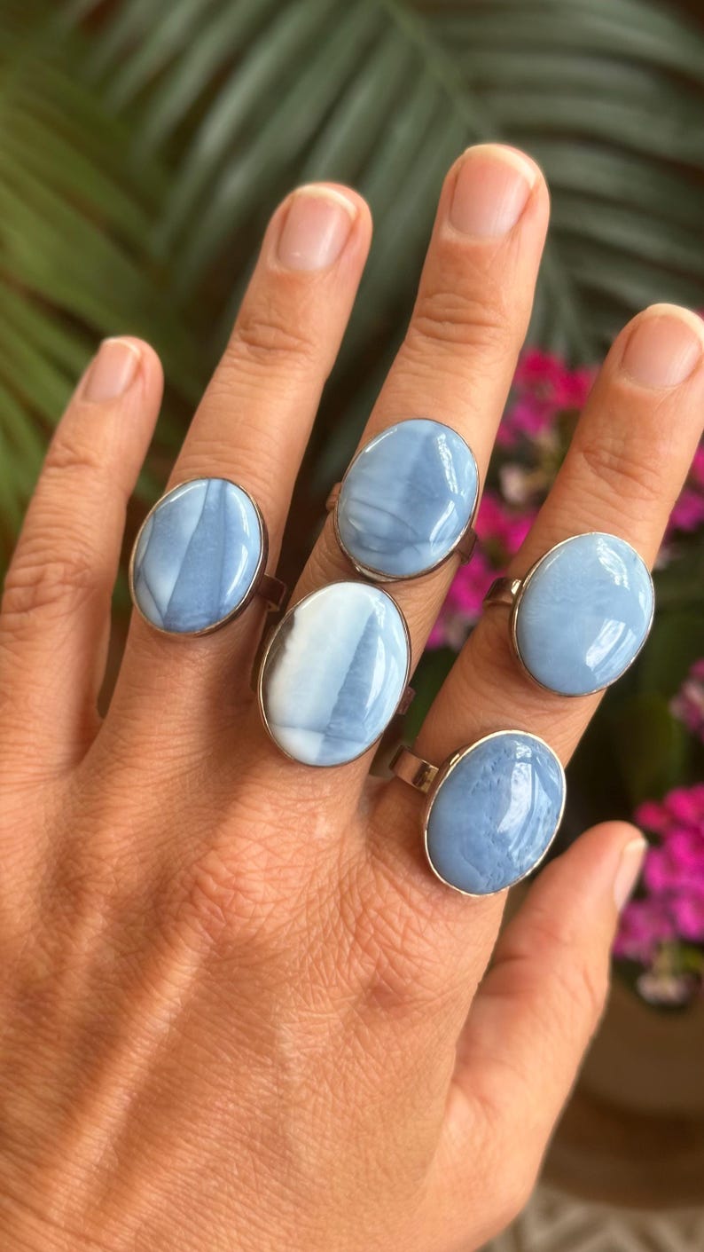 May include: Five silver rings with oval, light blue gemstones are displayed on a hand. The gemstones have a marbled appearance with white and blue swirls. The rings are set on a silver band.