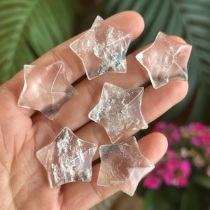 May include: Six clear quartz crystal star shapes. Each star has a five-point design and is translucent with internal inclusions. The stars are held in a hand, with a blurred green and pink background.