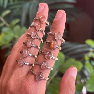 May include: A collection of ten silver rings, each featuring a single oval-shaped sunstone cabochon. The stones display varying shades of orange, peach, and translucent pink, set in simple metal bands worn across multiple fingers on one hand.