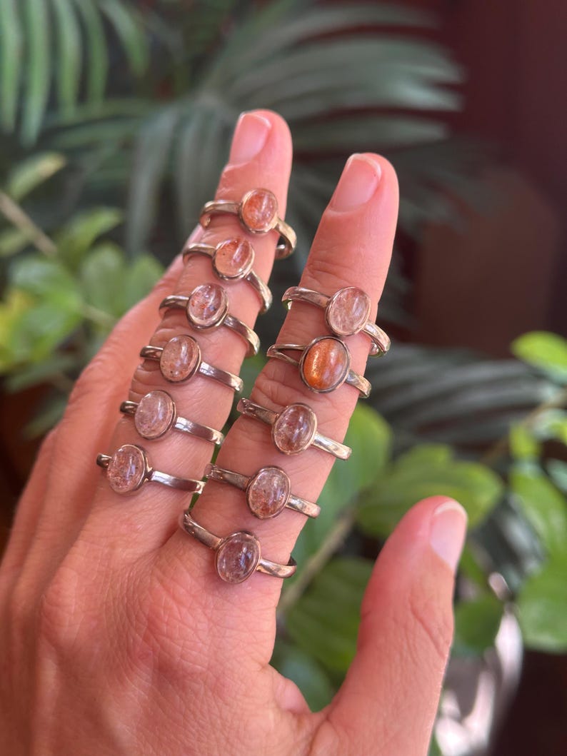 May include: A collection of ten silver rings, each featuring a single oval-shaped sunstone cabochon. The stones display varying shades of orange, peach, and translucent pink, set in simple metal bands worn across multiple fingers on one hand.