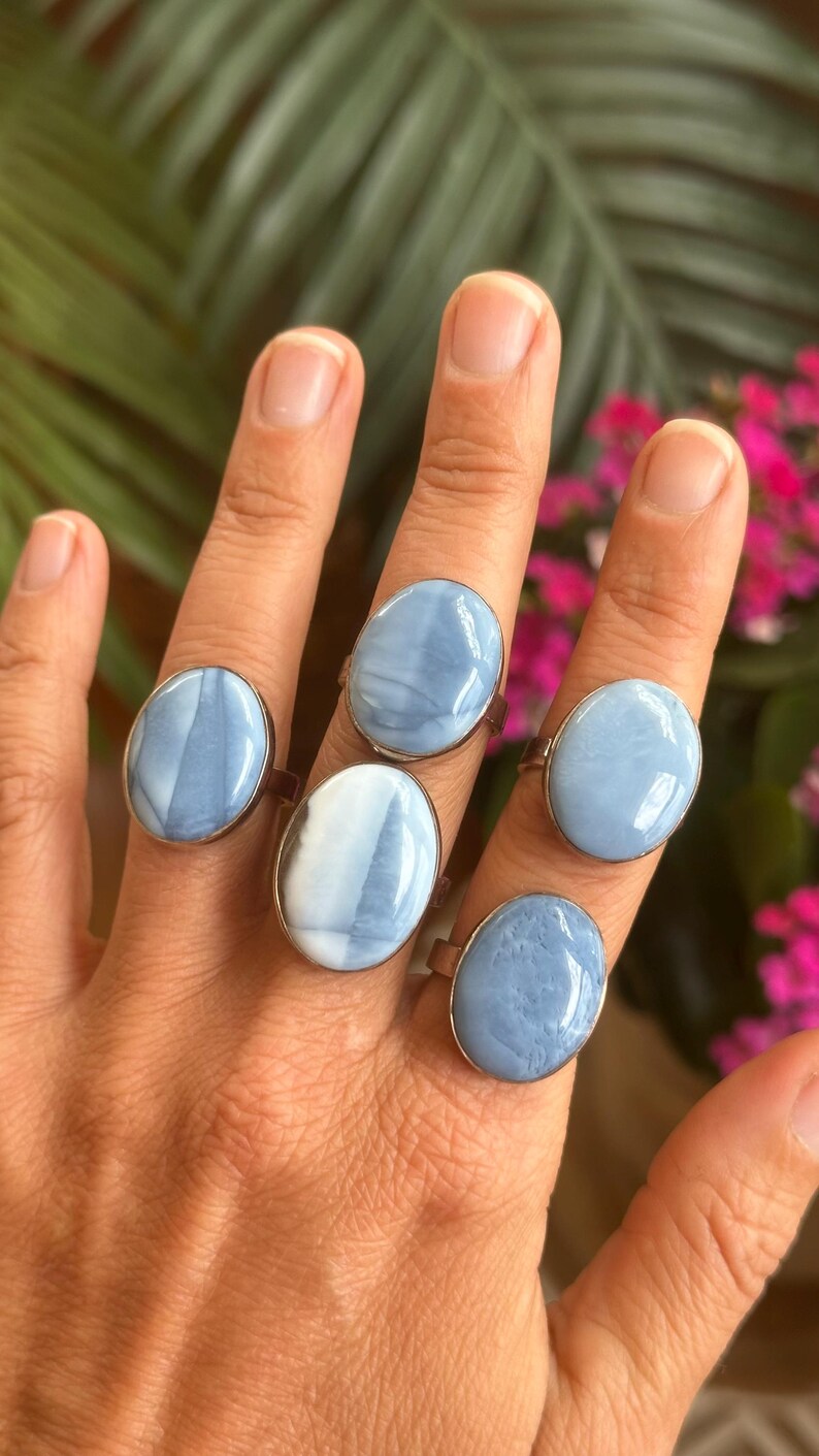 May include: Five oval-shaped rings with light blue and white stone settings are displayed on a hand. The rings have a silver-colored band. The background features green leaves and pink flowers.