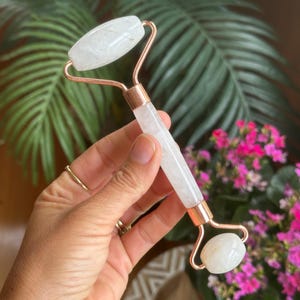 Puede incluir: Un rodillo facial de cuarzo blanco con detalles metálicos color cobre. El rodillo se sostiene en una mano, con un fondo de follaje verde y flores rosas. El rodillo está diseñado para el cuidado de la piel y el masaje facial.