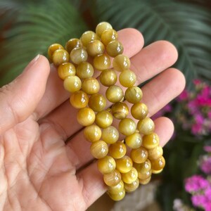 May include: A set of three golden-yellow tiger eye stone bracelets. Each bracelet is made of smooth, round beads. The bracelets are held in a hand, with a blurred background of green foliage and pink flowers.