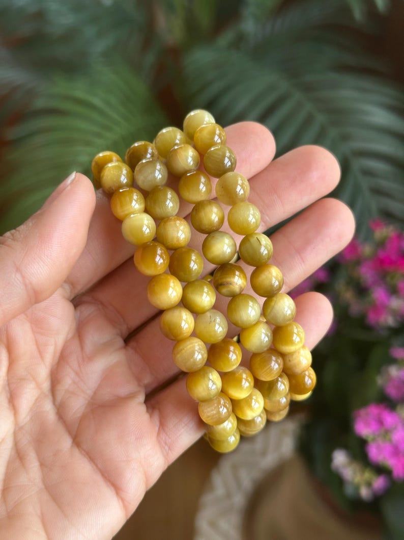 May include: A set of three golden-yellow tiger eye stone bracelets. Each bracelet is made of smooth, round beads. The bracelets are held in a hand, with a blurred background of green foliage and pink flowers.