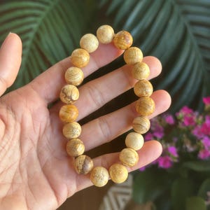 May include: A beaded bracelet featuring round, light brown wooden beads with natural grain patterns. The bracelet is held in a hand, showcasing the warm tones of the wood. The background includes green foliage and pink flowers.