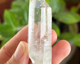 May include: A clear, elongated quartz crystal point with a hexagonal structure and natural facets. The mineral displays internal inclusions, varying levels of transparency, and a pointed termination at the top, showcasing its raw, unpolished form.
