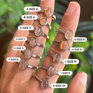 May include: A collection of eleven silver-toned rings featuring oval-shaped sunstone cabochons. Each ring is labeled with a letter from A to K and its corresponding US ring size, ranging from size 6 to size 10, clearly printed on white rectangular tags.