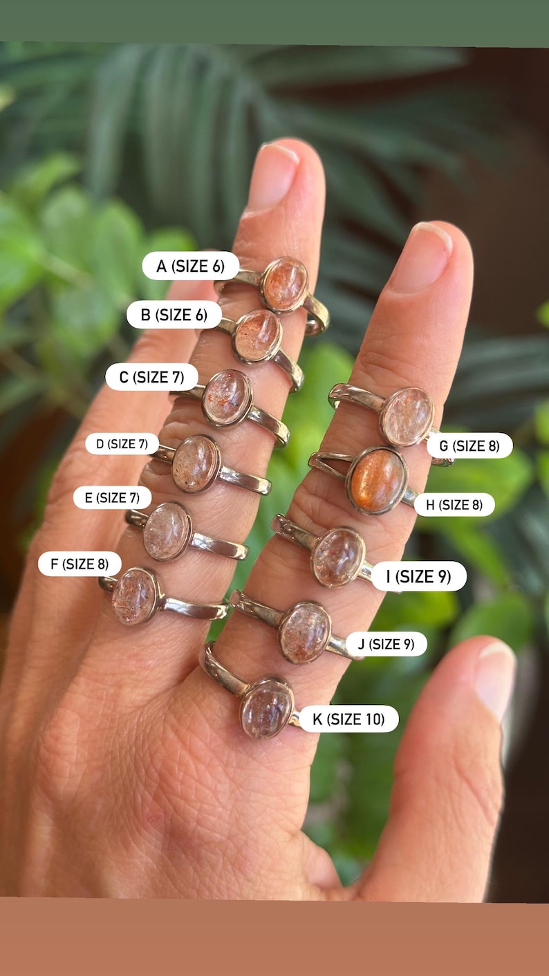 May include: A collection of eleven silver-toned rings featuring oval-shaped sunstone cabochons. Each ring is labeled with a letter from A to K and its corresponding US ring size, ranging from size 6 to size 10, clearly printed on white rectangular tags.