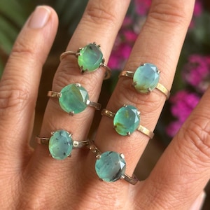May include: A hand displays six rings with faceted, oval, and round-cut gemstones in shades of green and blue. The rings have silver or gold bands. The background is blurred, with hints of green foliage and pink flowers.